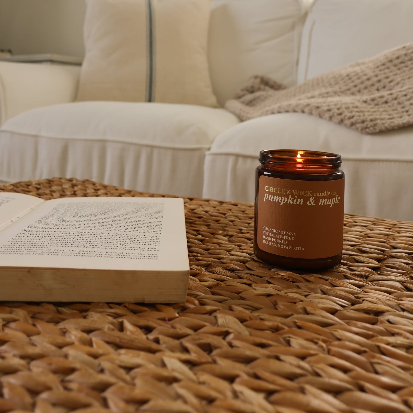 Candle labeled 'Pumpkin & Maple' on a woven table with an open book, in front of a white couch and framed picture.