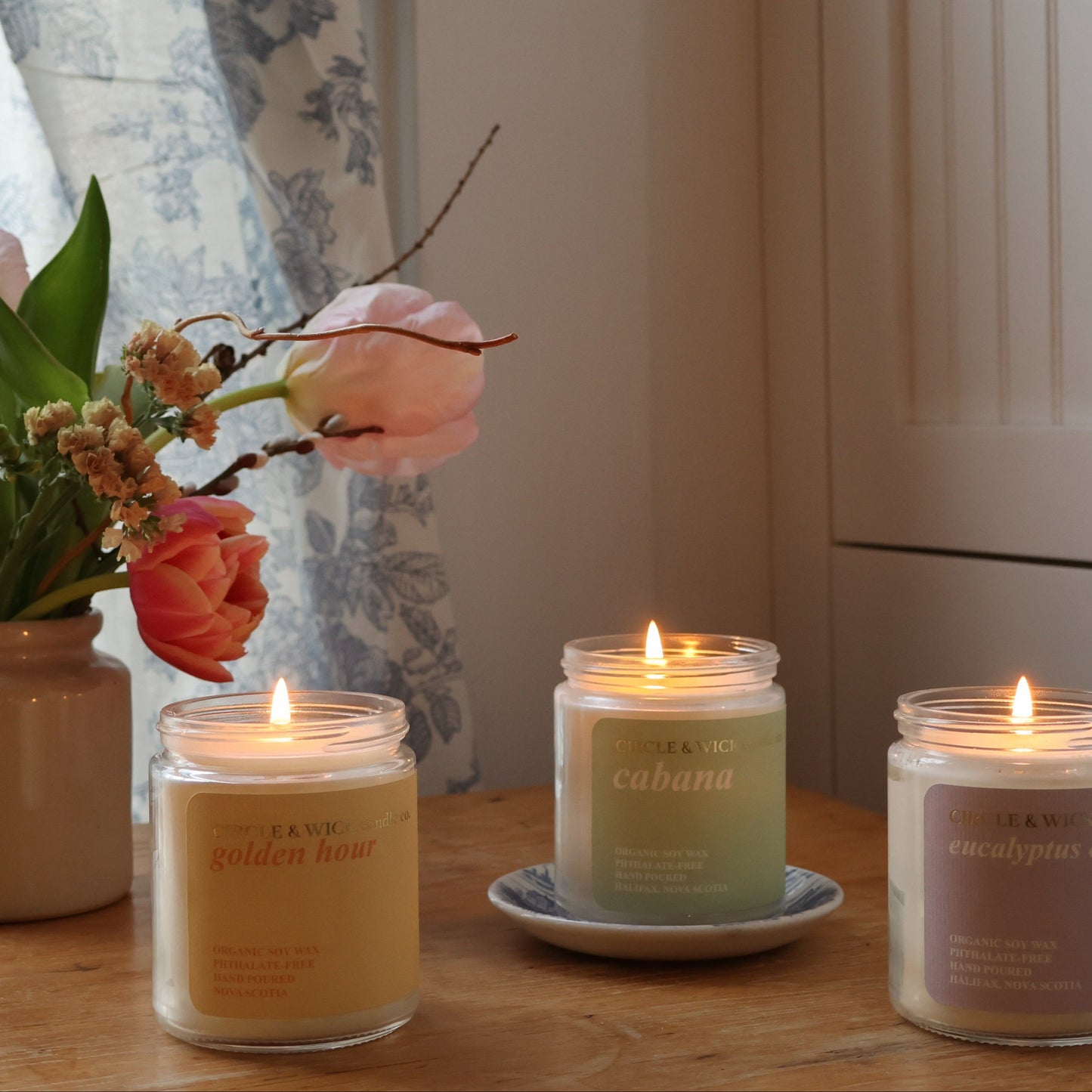 Three lit candles on a wooden surface with flowers in the background