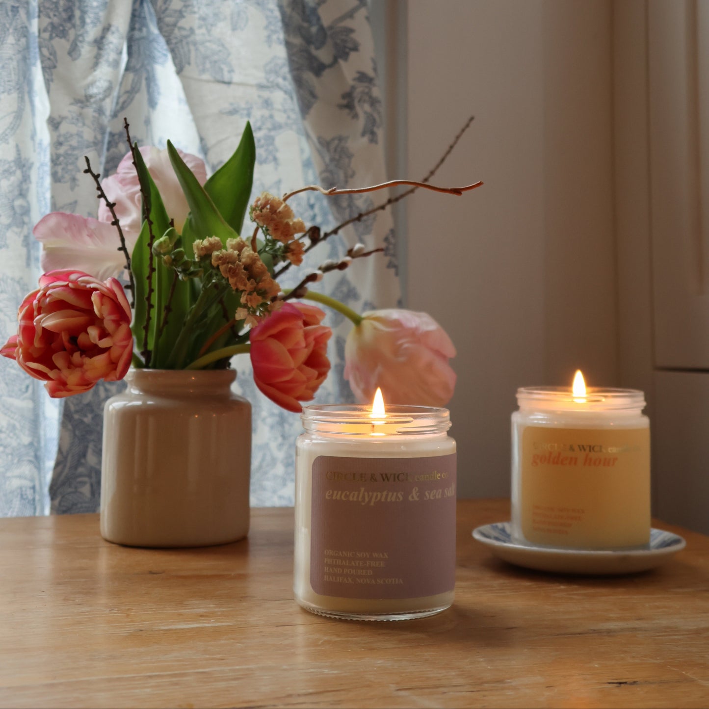 Two candles and a vase with flowers on a wooden surface near a window with floral curtains.